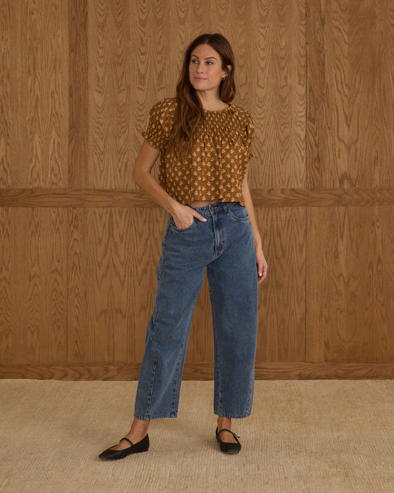 Woman wearing a patterned top and blue jeans standing against a wooden paneled wall.